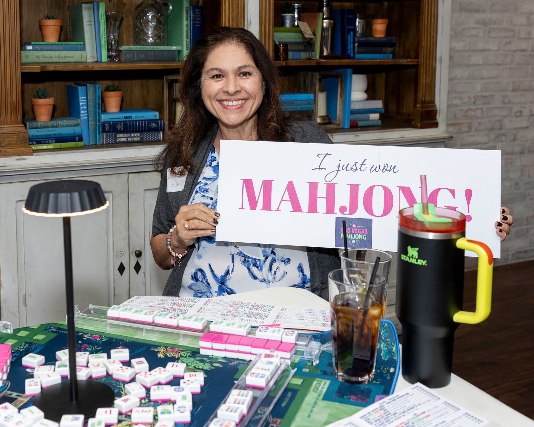 A smiling woman holding a sign that reads I just won Mahjong, with the Las Vegas Mahjong logo, seated at a table with her tiles and a drink