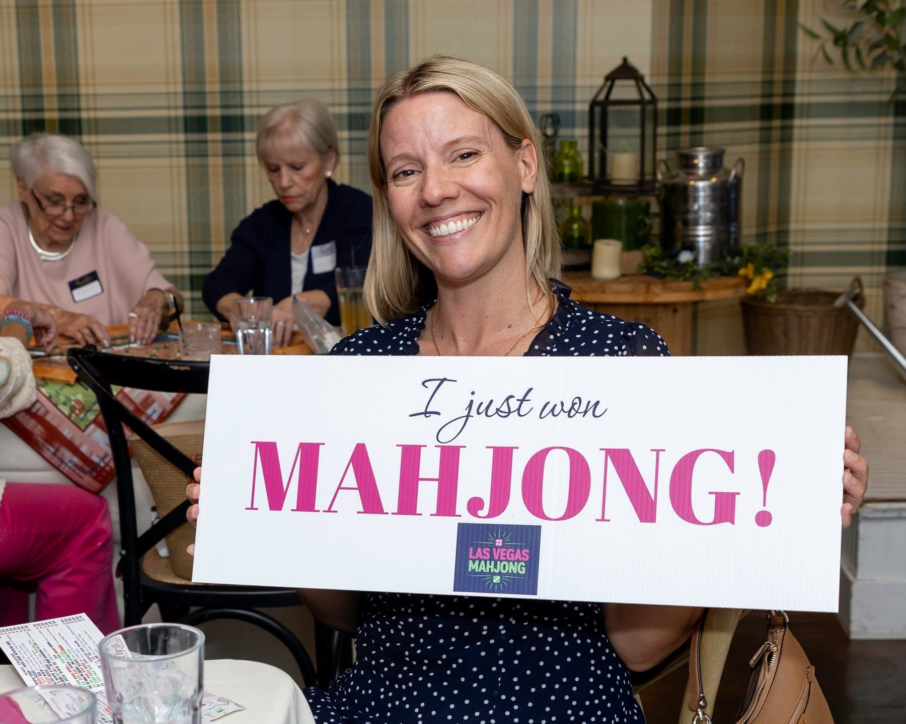 A smiling woman holding a sign that reads I just won Mahjong, with the Las Vegas Mahjong logo, while other guests keep playing behind her