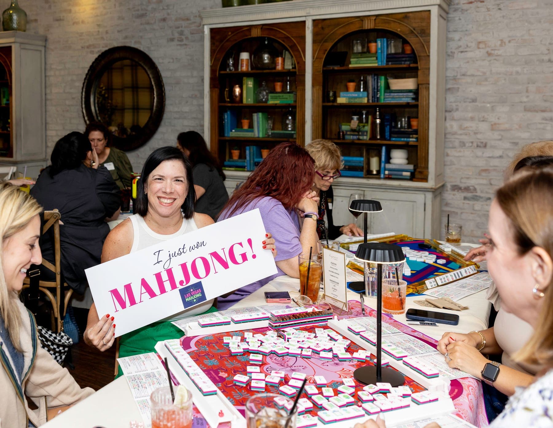 A smiling woman holding a sign that reads I just won Mahjong, with the Las Vegas Mahjong logo, while other guests play mahjong at nearby tables