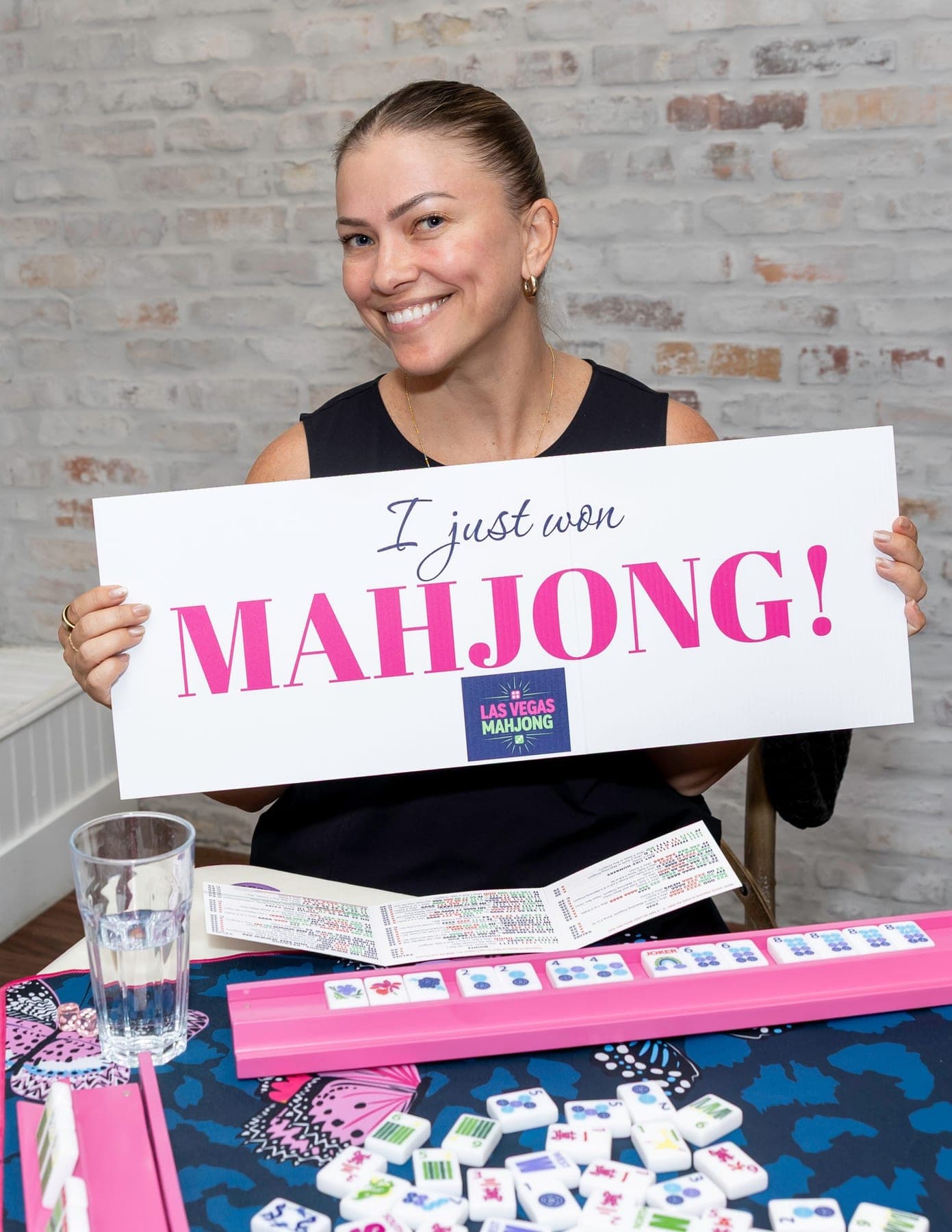 A smiling woman holding a sign that reads I just won Mahjong, with the Las Vegas Mahjong logo, seated at a table with her mahjong card and tiles
