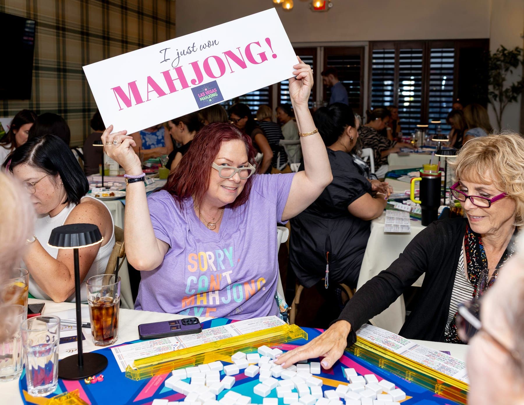 A smiling woman holding a sign overhead that reads I just won Mahjong, with the Las Vegas Mahjong logo, in a room full of other players