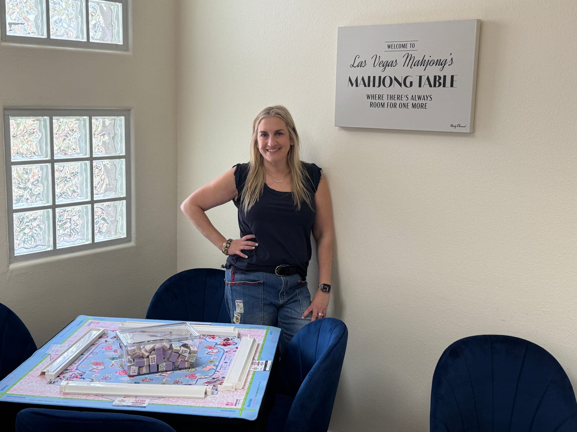 Shauna inside the Las Vegas Mahjong Studio beside a mahjong table, under a sign reading Welcome to Las Vegas Mahjong's Mahjong Table