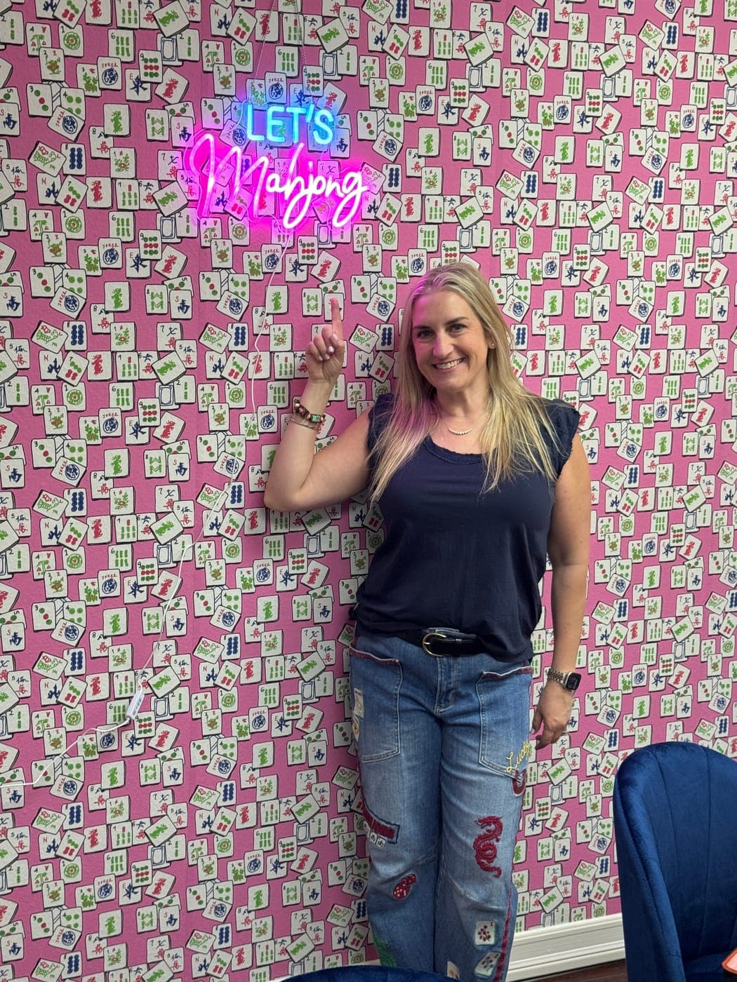 Shauna inside the Las Vegas Mahjong Studio, pointing to a neon sign reading Let's Mahjong on the pink tile-patterned wall